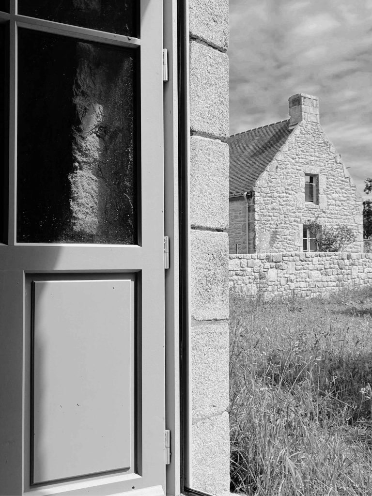 Vue depuis une menuiserie bois vers une maison en pierres dans un hameau en zone protégée à Carnac